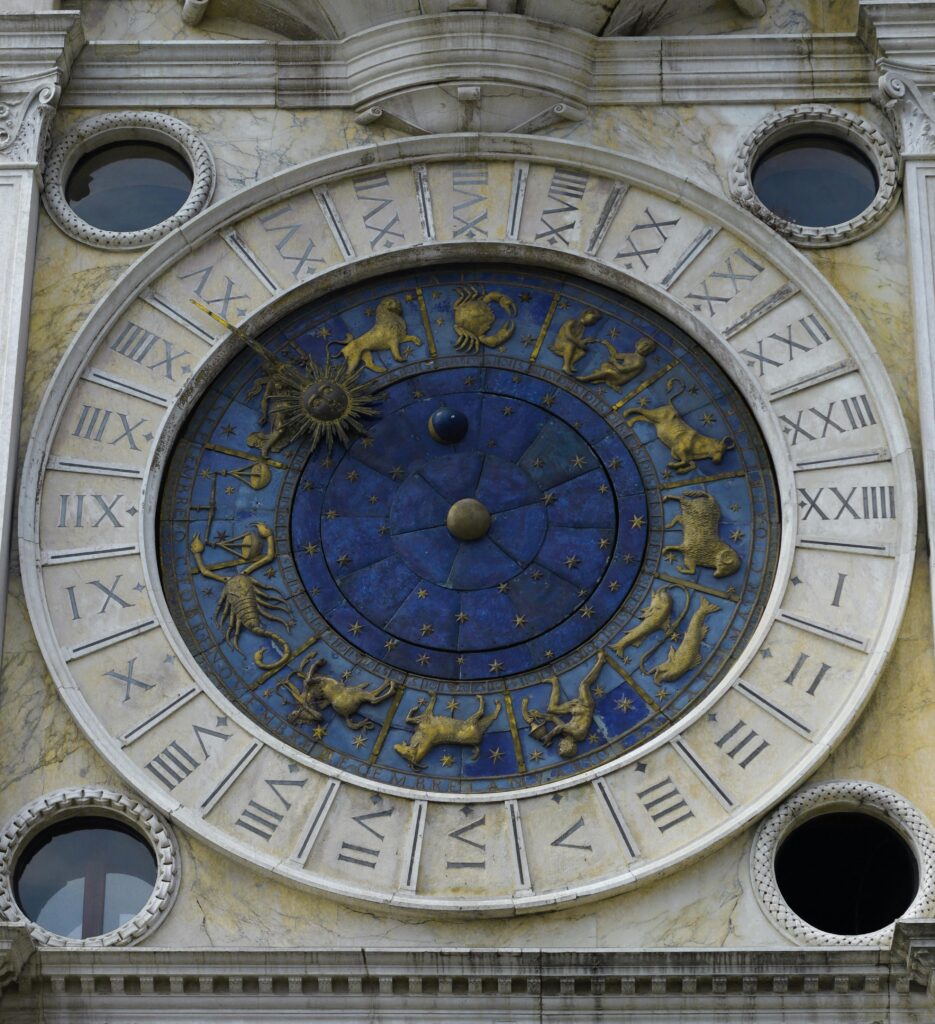 Détail d'une horloge astronomique ancienne montrant les signes du zodiaque sculptés en doré sur un cadran bleu étoilé, entouré de chiffres romains.