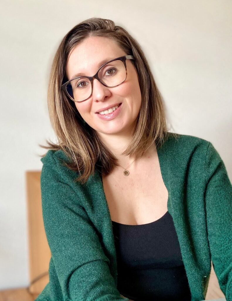 Smiling woman with shoulder-length brown hair and black-framed glasses wearing a green cardigan.