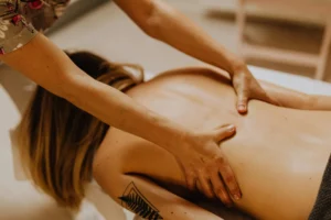 Massage therapist kneads a person’s back while they lie face down on a table; a tattoo is visible on the upper leg/hip area.