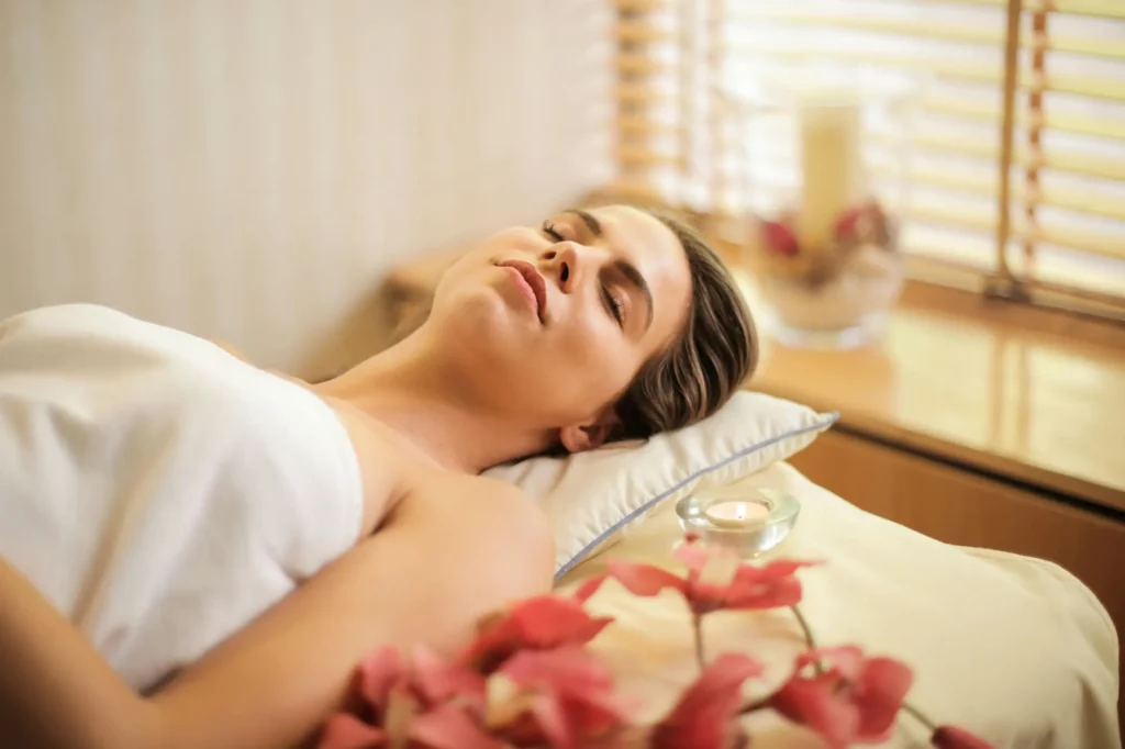 Illustration_femme_paisible | Espace Sélènes Une femme allongée, les yeux fermés, se relaxant dans une ambiance de spa avec des fleurs et une bougie.