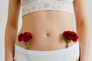 Plan moyen du ventre d'une femme debout, portant des sous-vêtements blancs. Deux œillets rouges sont glissés de chaque côté de la taille, au niveau des hanches, pour symboliser les ovaires.